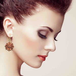 4 of 7: Woman with hair styled up wearing copper sunburst flower dangle earrings. The polished copper floral charm features layered petal detailing in a radiant sunburst pattern and hangs from a brass French ear wire. Shown in a soft profile pose with eyes down, the image highlights the warm copper tone, delicate movement, and elegant drop length of the handcrafted earring against a light neutral background.