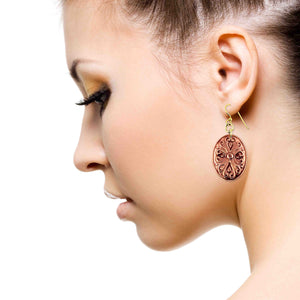 6 of 7: Close-up side profile of a woman wearing a handcrafted copper Victorian oval dangle earring with ornate scrollwork, suspended from a brass French ear wire, highlighting the warm polished copper tone against her skin and soft neutral makeup on a clean white background.