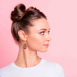 2 of 7: Woman wearing handcrafted copper Victorian oval dangle earring with detailed scrollwork, styled with hair in a neat bun, shown in side profile against a soft pink background, highlighting the warm copper tones and elegant drop design.