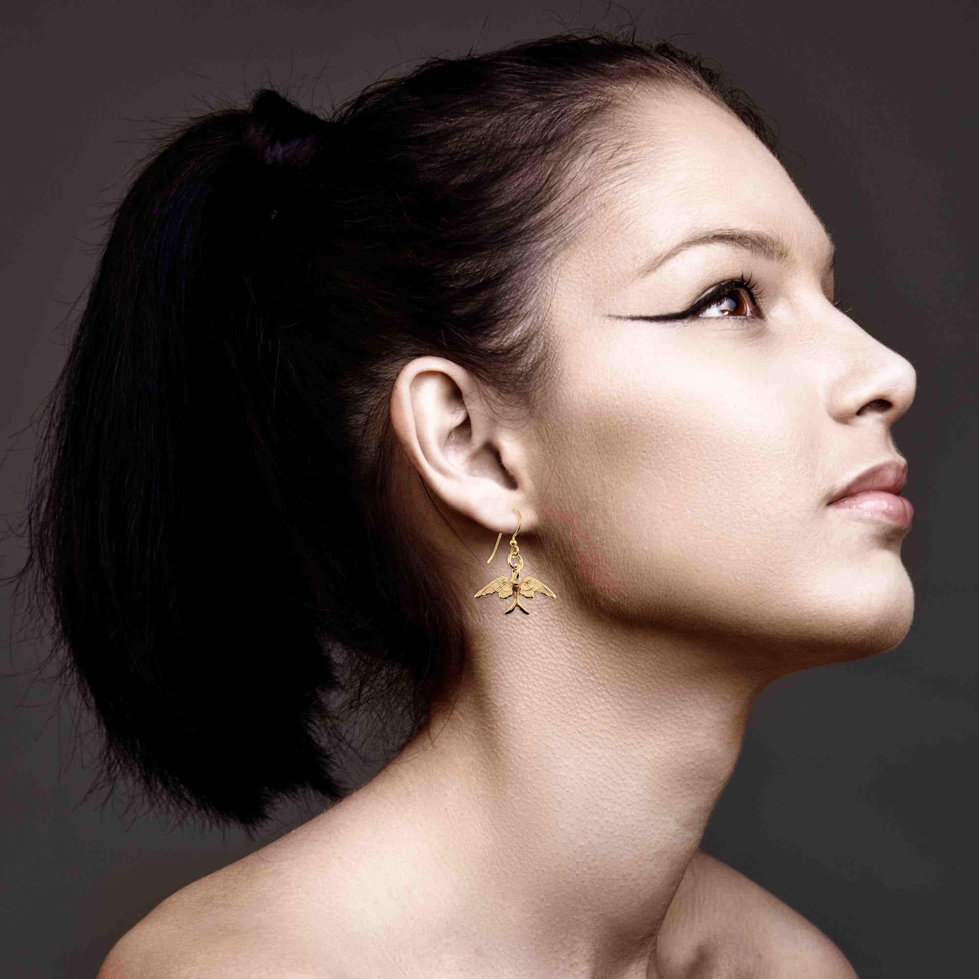 Woman wearing Dainty Nu Gold Brass Swallow Earrings against a dark background