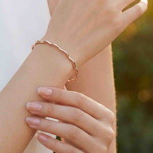 6 of 9: Close up of a woman’s wrist wearing a slim polished copper cuff bracelet with softly wavy, scalloped edges. The open bracelet wraps lightly around the wrist and reflects warm natural light. Her other hand rests gently below, showing pale pink manicured nails, while a softly blurred outdoor background gives the image a bright, elegant feel.