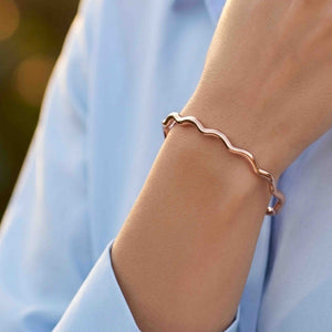 2 of 9: Close up of a wrist wearing a slim polished copper cuff bracelet with softly wavy, scalloped edges. The open cuff sits just above the wrist bone and has a smooth reflective finish that catches the light. The bracelet is styled with a light blue long sleeve top, with a softly blurred outdoor background that gives the image a warm natural feel.