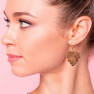 2 of 7: A woman wearing a handcrafted copper grape leaf dangle earring with a brass French hook. The earring features detailed leaf vein patterns and hangs just below the earlobe. The background is a soft pink, highlighting the warm tones of the copper and the natural elegance of the jewelry.