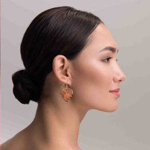 4 of 7: A woman with dark hair styled in a low bun is wearing a handcrafted copper grape leaf dangle earring with a brass French hook. The earring features detailed veining that mimics a real grape leaf and hangs just below her earlobe. The neutral gray background and soft lighting highlight the warm tone and texture of the copper jewelry.