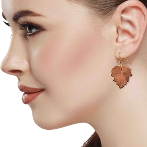 6 of 7: A close-up side profile of a woman wearing a handcrafted copper grape leaf dangle earring with a brass French hook. The earring features detailed leaf veining and hangs just below the earlobe. The background is white, drawing focus to the warm tones of the copper jewelry and the model's natural makeup.
