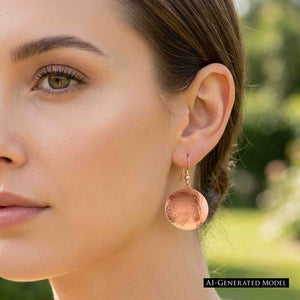 2 of 7: Woman with brown hair pulled back wearing a Hammered Copper Disc Earring outdoors in natural light. The round copper disc hangs below the ear and displays its hand hammered texture, polished finish, and noticeable size when worn. AI generated model image.