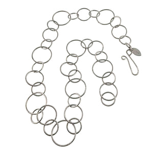 1 of 8: Full hammered stainless steel chain link necklace arranged on a white background with its handmade hook clasp and John S. Brana metal tag visible. The necklace features repeating round links in three sizes, with a polished silver tone finish and hammered texture.