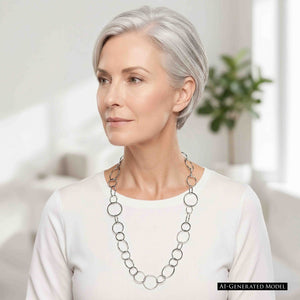 2 of 8: AI generated mature model with short silver hair wearing a long hammered stainless steel chain link necklace over a simple white top. The mixed size polished silver tone links stand out clearly against the light clothing in a bright indoor setting.