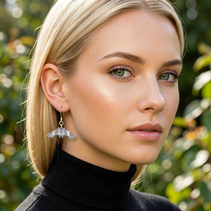 2 of 8: Blonde woman wearing a polished aluminum bat dangle earring with a gold tone ear wire and a black turtleneck. The spread wing bat hangs below her ear and stands out against her straight blonde hair and the softly blurred outdoor background.
