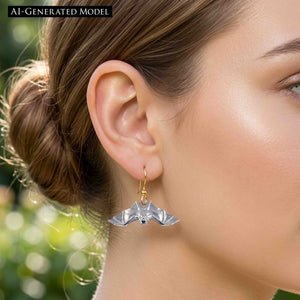 6 of 8: Close view of a blonde woman wearing a silver tone bat dangle earring with a gold tone French ear wire. The polished bat has spread wings and reflective details, with the ear and jewelry shown against blonde hair and a softly blurred green background.  AI Generated Model.