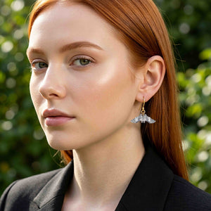 6 of 8: Close portrait of a red haired woman wearing a polished aluminum bat dangle earring with a gold tone ear wire. Her hair is tucked behind her ear, and she wears a black blazer against a softly blurred green outdoor background.