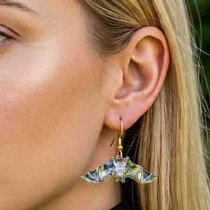 7 of 8: Close view of a blonde woman wearing a silver tone bat dangle earring with a gold tone French ear wire. The polished bat has spread wings and reflective details, with the ear and jewelry shown against blonde hair and a softly blurred green background.