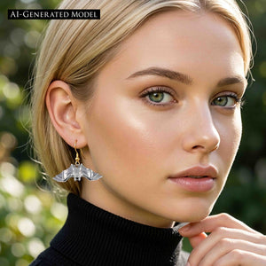 2 of 8: Blonde woman wearing a polished aluminum bat dangle earring with a gold tone ear wire and a black turtleneck. The spread wing bat hangs below her ear and stands out against her straight blonde hair and the softly blurred outdoor background.  AI Generated Model.