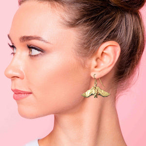 2 of 7: Close-up of a woman wearing Hanging Bat Nu Gold Brass Dangle Earrings against a pink background