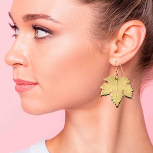 2 of 7: Close-up of a woman wearing Large Maple Leaf Nu Gold Brass Dangle Earrings against a pink background