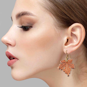 6 of 7: Close-up of a woman wearing a Medium Maple Leaf Copper Dangle Earring against a gray background
