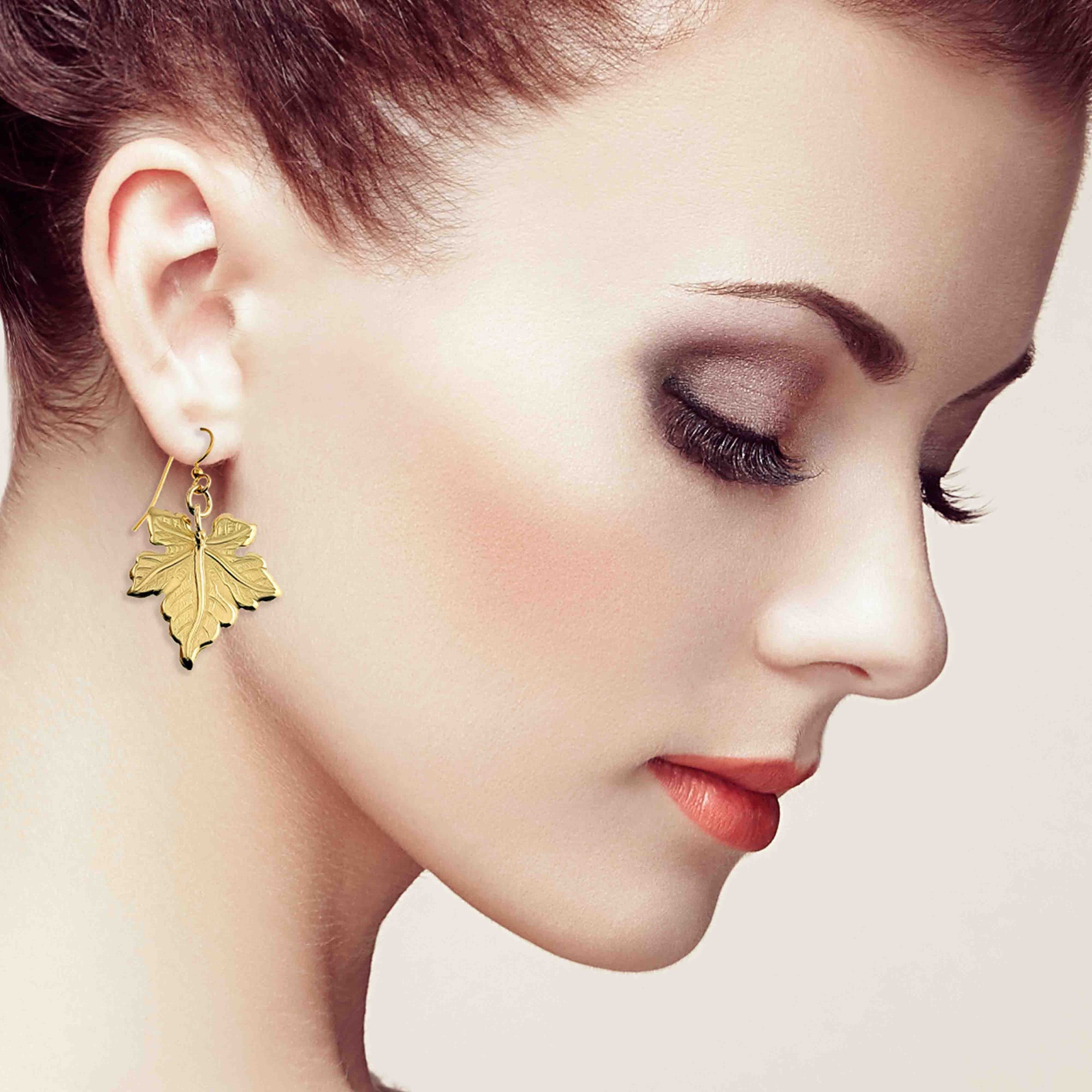 Close-up of a woman wearing a Medium Maple Leaf Nu Gold Brass Dangle Earring on a light background
