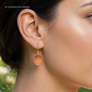 6 of 7: Close view of an AI generated female model wearing a Mini Hammered Copper Oval Disc Earring with a brass French ear wire. The polished copper oval hangs just below the earlobe, showing its warm reddish color and hand hammered texture against dark hair and a softly blurred green outdoor background.