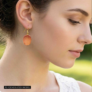 2 of 7: Side profile of an AI generated female model wearing a Mini Hammered Copper Oval Disc Earring outdoors. The small polished copper oval hangs from a brass French ear wire and catches the light, clearly showing the hand hammered surface. The model wears her dark hair pulled back with a white top against a soft green background.