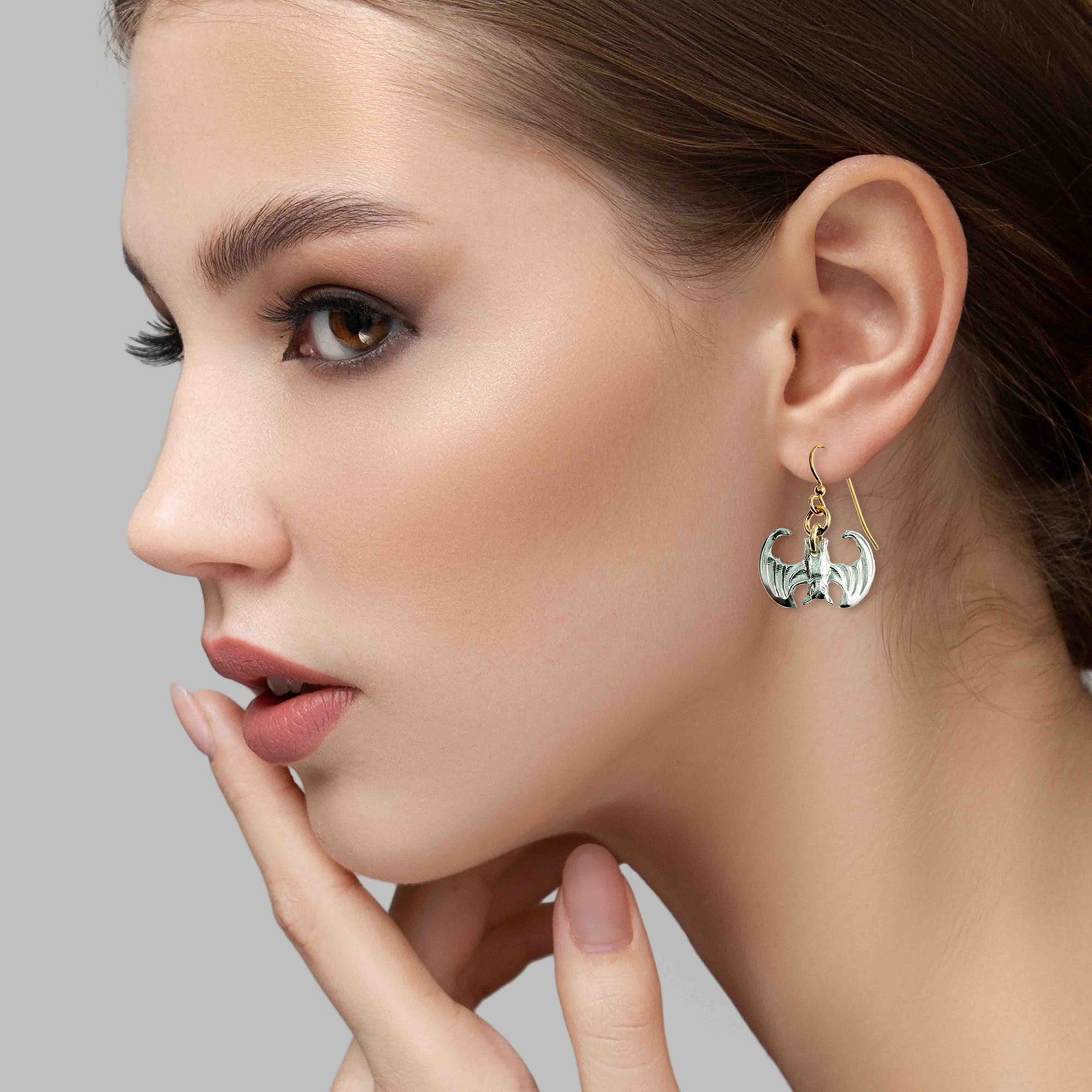 Close-up of a woman wearing Mini Vampire Bat Aluminum Dangle Earrings  on a gray background