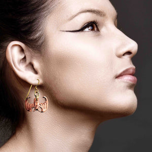 6 of 7: Close-up side profile of a woman wearing a handcrafted copper vampire bat dangle earring with brass French ear wire, paired with bold, winged eyeliner, against a dark background.