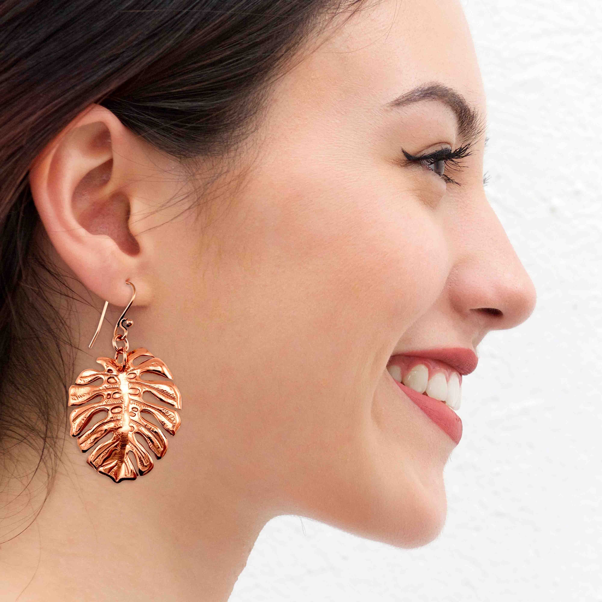 Side profile of a smiling woman with dark hair pulled back, wearing a shiny copper monstera leaf dangle earring on a gold tone French hook, the detailed cutout leaf hanging just below her earlobe against a textured white background.