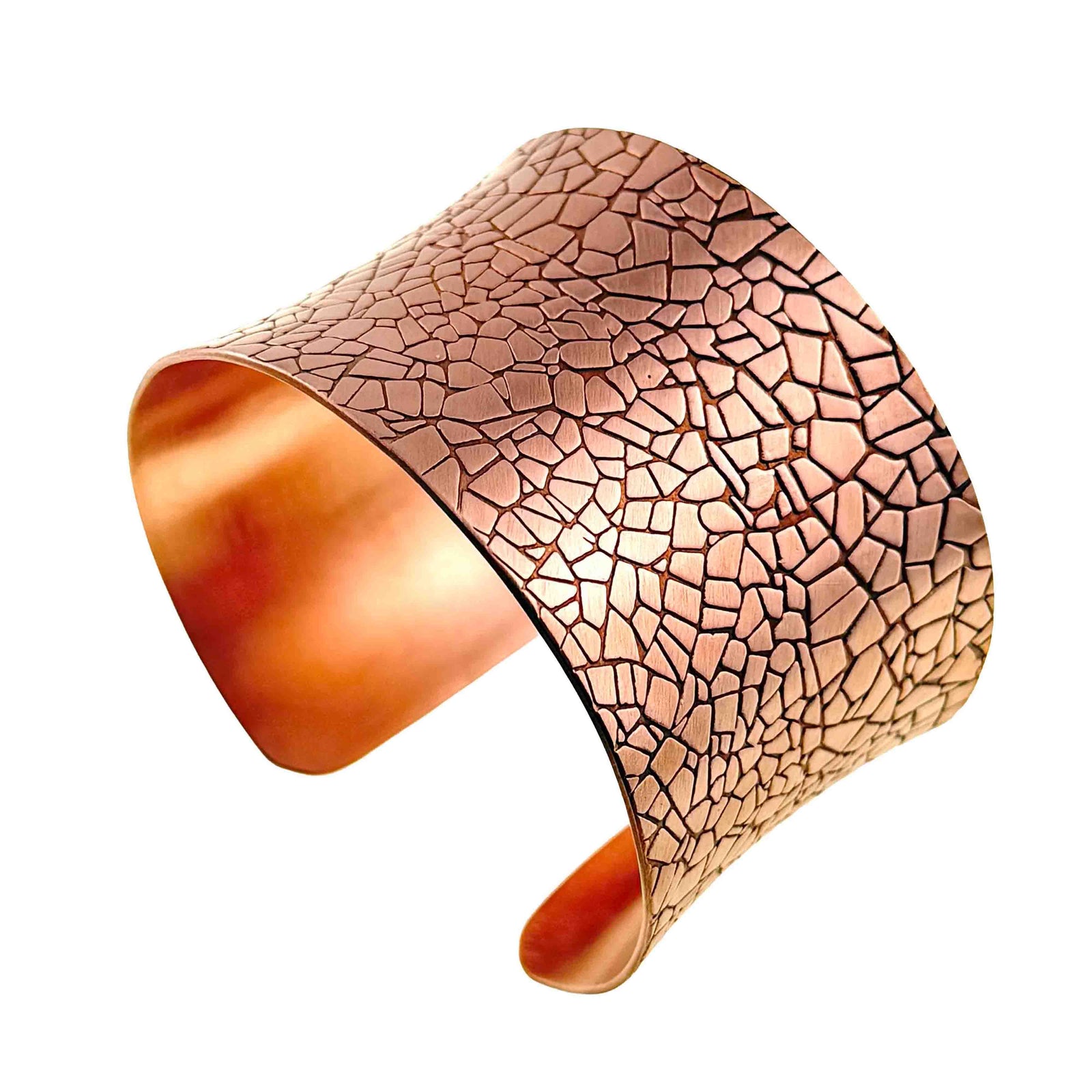 Angled product view of a wide solid copper cuff bracelet on a white background. The anticlastic shape curves inward at the center and flares gently at the edges. An embossed mosaic pattern covers the brushed outer surface, while the open interior shows smooth polished copper.