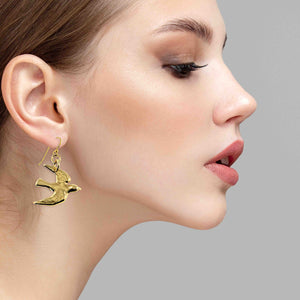 6 of 7: Close-up of a woman wearing a Nu Gold Brass Flying Swallow Earring against a gray background
