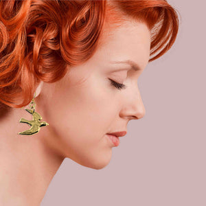 4 of 7: Woman with red hair wearing a Nu Gold Brass Flying Swallow Earring against a pink background