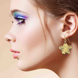 6 of 7: Close-up of a woman's profile wearing a Nu Gold Brass Hibiscus Flower Dangle Earring  against a beige background