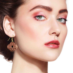4 of 7: Close-up portrait of a woman wearing ornate clover-shaped copper drop earrings with detailed scrollwork and a small center cutout. The warm copper tone contrasts softly with her skin, while gold tone brass French ear wires add a subtle shine. The image highlights how the earrings hang just below the earlobe and complement natural makeup with soft pink tones.