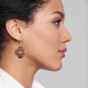 2 of 7: Side profile of a woman wearing ornate clover-shaped copper drop earrings with detailed scrollwork and a small center cutout. The warm copper design hangs from gold tone brass French ear wires, adding a soft glow against her skin. The image highlights how the earrings sit just below the earlobe and complement a clean, natural look.