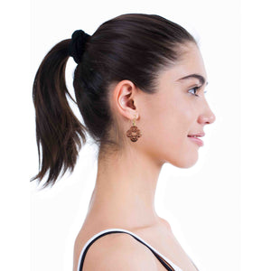 6 of 7: Side profile of a woman with dark hair in a ponytail wearing ornate clover-shaped copper drop earrings. The earrings feature detailed scrollwork and a small center cutout, hanging from gold tone brass French ear wires. The image shows how the earrings sit just below the earlobe and add a warm, polished accent to a clean, casual look.