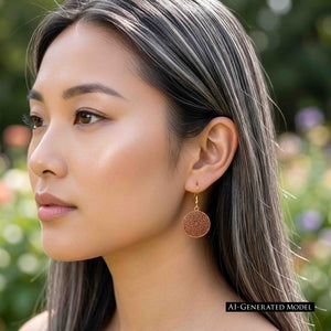 4 of 8: AI Generated Model wearing a Small Copper Mandala Disc Earring outdoors. The round copper drop earring hangs from a brass French ear wire and shows a detailed swirling mandala pattern against the model's dark hair, with a softly blurred green garden background.