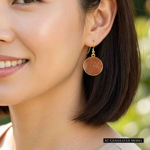 6 of 8: AI Generated Model wearing a Small Copper Mandala Disc Earring outdoors. The round copper earring hangs from a brass French ear wire beside the model's short dark hair. Its swirling mandala pattern is clearly visible, with warm copper tones set against a softly blurred green background.