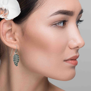 2 of 8: A woman with smooth skin and dark hair styled in a low bun wears a white orchid tucked above her ear. She is shown in profile against a soft gray background. She is wearing a small aluminum feather dangle earring with detailed texture and a stainless steel French ear wire. The earring hangs just below her earlobe, adding a light, natural touch to her look.