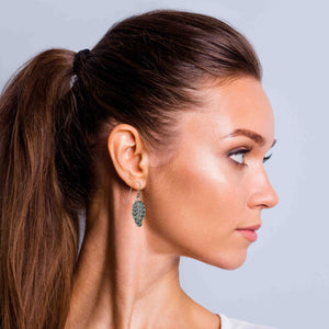 4 of 8: A woman with long brown hair pulled back in a high ponytail is shown in profile against a light gray background. She is wearing a small aluminum feather dangle earring that hangs just below her earlobe. The earring has textured detailing and a stainless steel French ear wire. The lighting highlights her skin tone and the earring’s soft metallic shine.