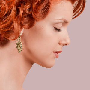 6 of 7: Woman with red hair wearing John S. Brana Small Feather Bronze Dangle Earrings, highlighting the detailed feather design and warm bronze tone against a soft pink background.