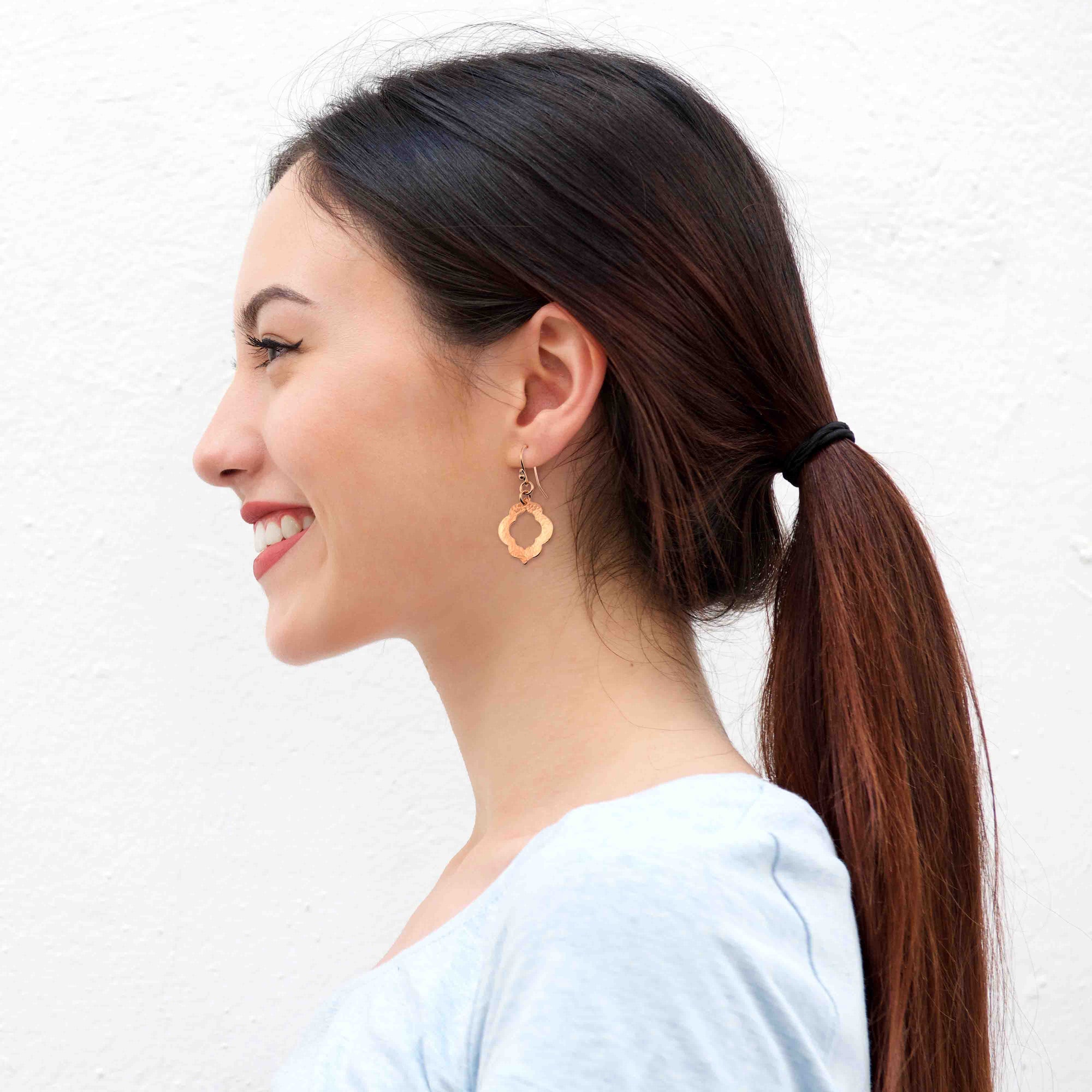 A Smiling Woman Wearing Small Hammered Copper Pierced Quatrefoil Dangle Earrings
