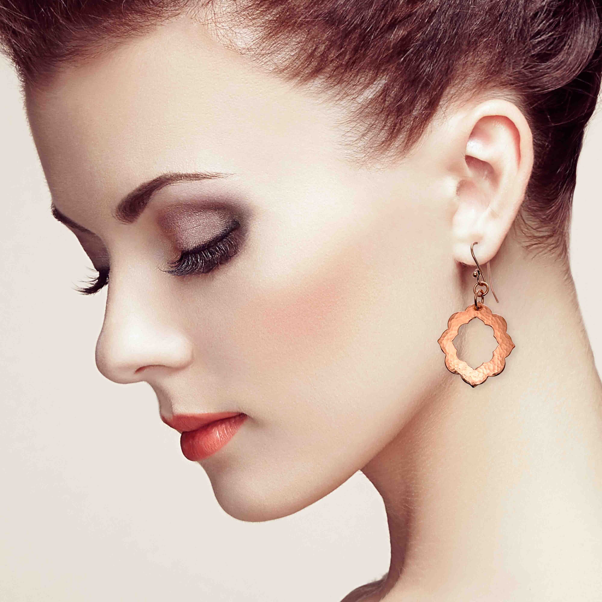 Close-up of a woman wearing Small Pierced Hammered Copper Arabesque Flower Earrings against a neutral background