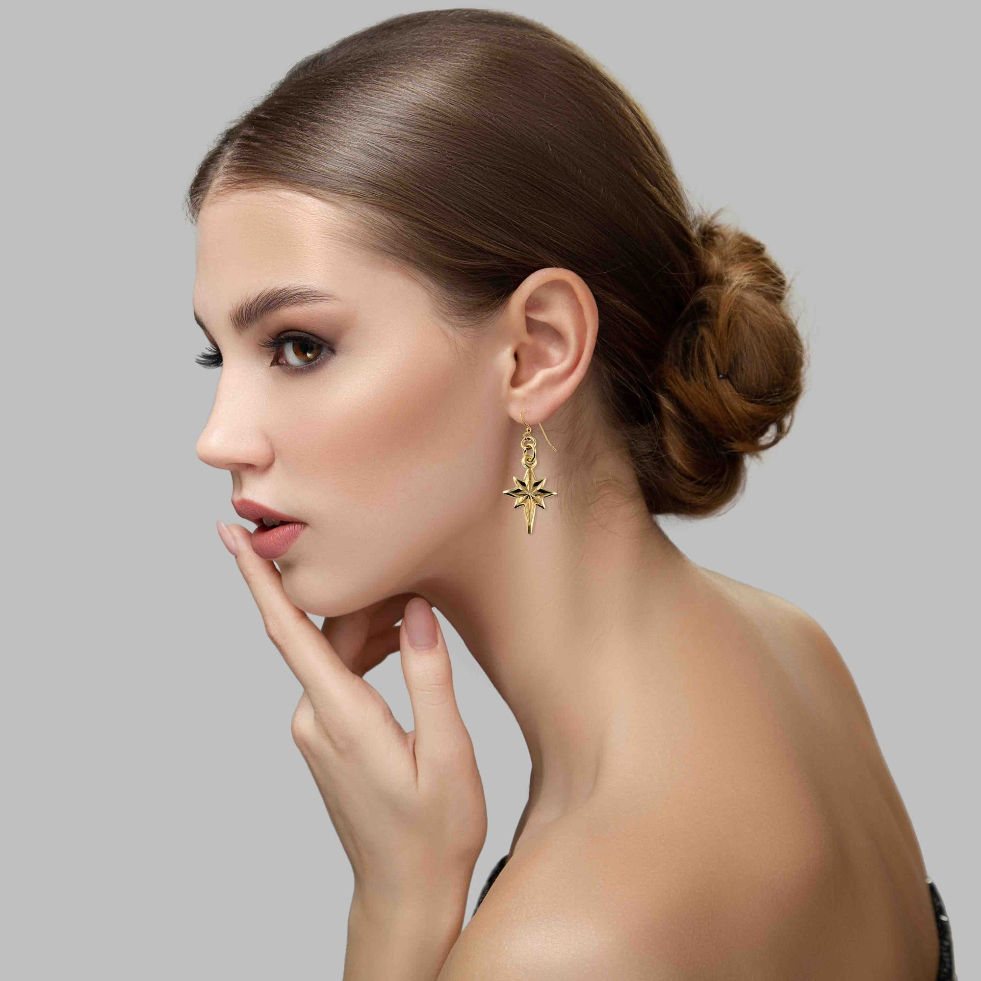 Woman wearing Star of Bethlehem Nu Gold Brass Dangle Earrings against a gray background