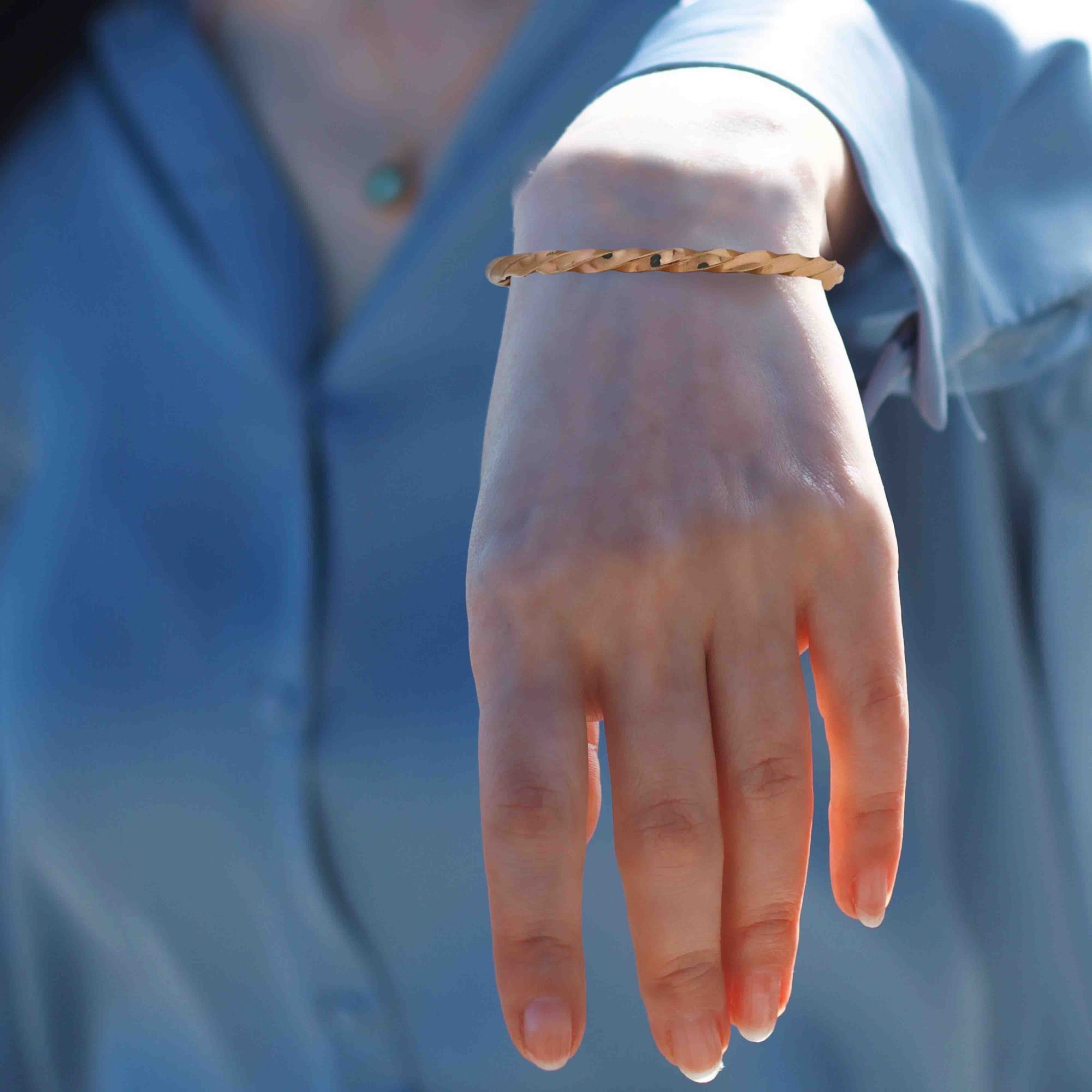 AI-generated model wearing a slim twisted solid copper cuff bracelet on the wrist. The handmade open cuff has a warm rose-copper tone, narrow rope-like texture, rounded ends, and reflective polished facets. The wrist is shown with the hand relaxed downward, framed by a soft blue shirt and blurred blue background, highlighting the bracelet’s delicate everyday style.