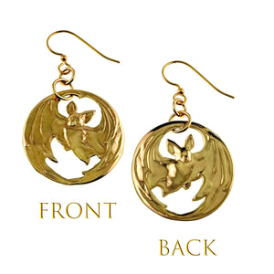3 of 7: Front and back view of gold-tone brass disc earrings featuring a die-struck bat with outstretched wings and cutout details. The front shows a more matte textured finish, while the back appears smoother and more polished. Each earring hangs from a brass French hook with small connecting rings, highlighting the dimensional bat design within the circular frame.