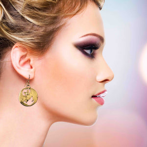 6 of 7: Side profile of a woman with blonde hair styled up, wearing gold-tone brass disc earrings featuring a detailed bat with outstretched wings and open cutout accents. The earring hangs from a French hook and sits just below the earlobe. Her soft makeup and neutral background highlight the warm metallic shine and bold, playful design.