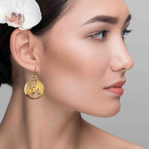 2 of 7: Side profile of a woman wearing gold-tone brass disc earrings with a detailed bat design. The die-struck bat features spread wings with open cutout areas, creating depth and light contrast. The earring hangs from a French hook and rests just below the earlobe, adding a warm metallic accent against her smooth skin and pulled-back dark hair with a white flower accessory.