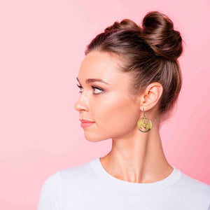 5 of 7: Side profile of a woman with her hair styled in a high bun, wearing gold-tone brass disc earrings featuring a detailed bat with outstretched wings and cutout accents. The earring hangs from a French hook and rests just below the earlobe. Set against a soft pink background, the warm metallic finish stands out, adding a playful yet bold touch to her clean, minimal look.