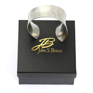 7 of 7: Anticlastic Tapered Linen Aluminum Bangle Bracelet on top of a Black Gift Box with a Gold Logo
