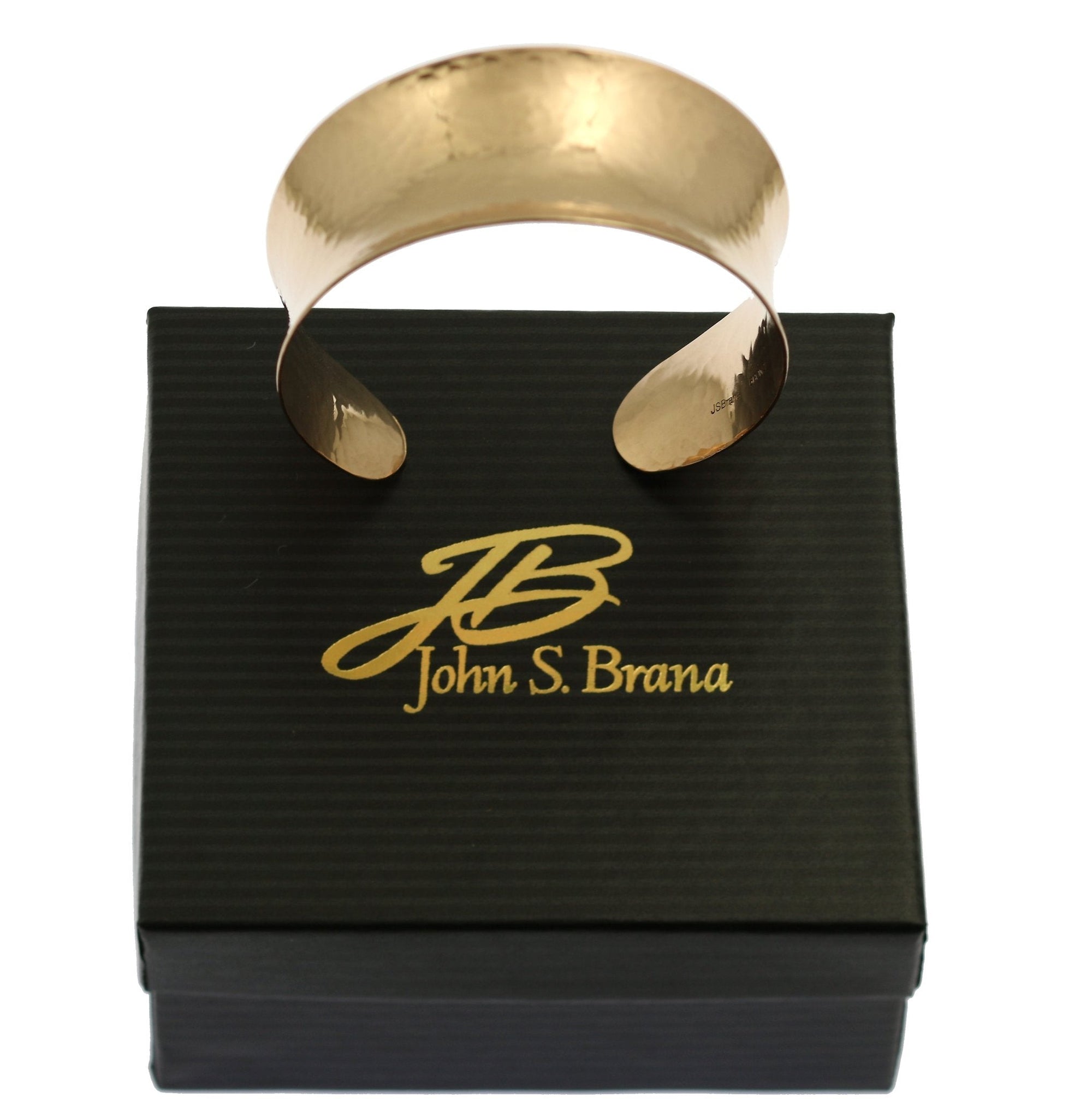 A handcrafted copper cuff bracelet with a hammered finish, displayed resting on top of a black gift box. The bracelet has a gently flared shape with smooth, rounded edges and an open-ended design for easy adjustment. Its polished surface catches the light, showing off the subtle hammered texture. The gift box features the John S. Brana logo in gold lettering with a stylized “JB” above the designer's full name. The setup suggests a ready-to-gift presentation, ideal for special occasions. Photographed against a plain white background.